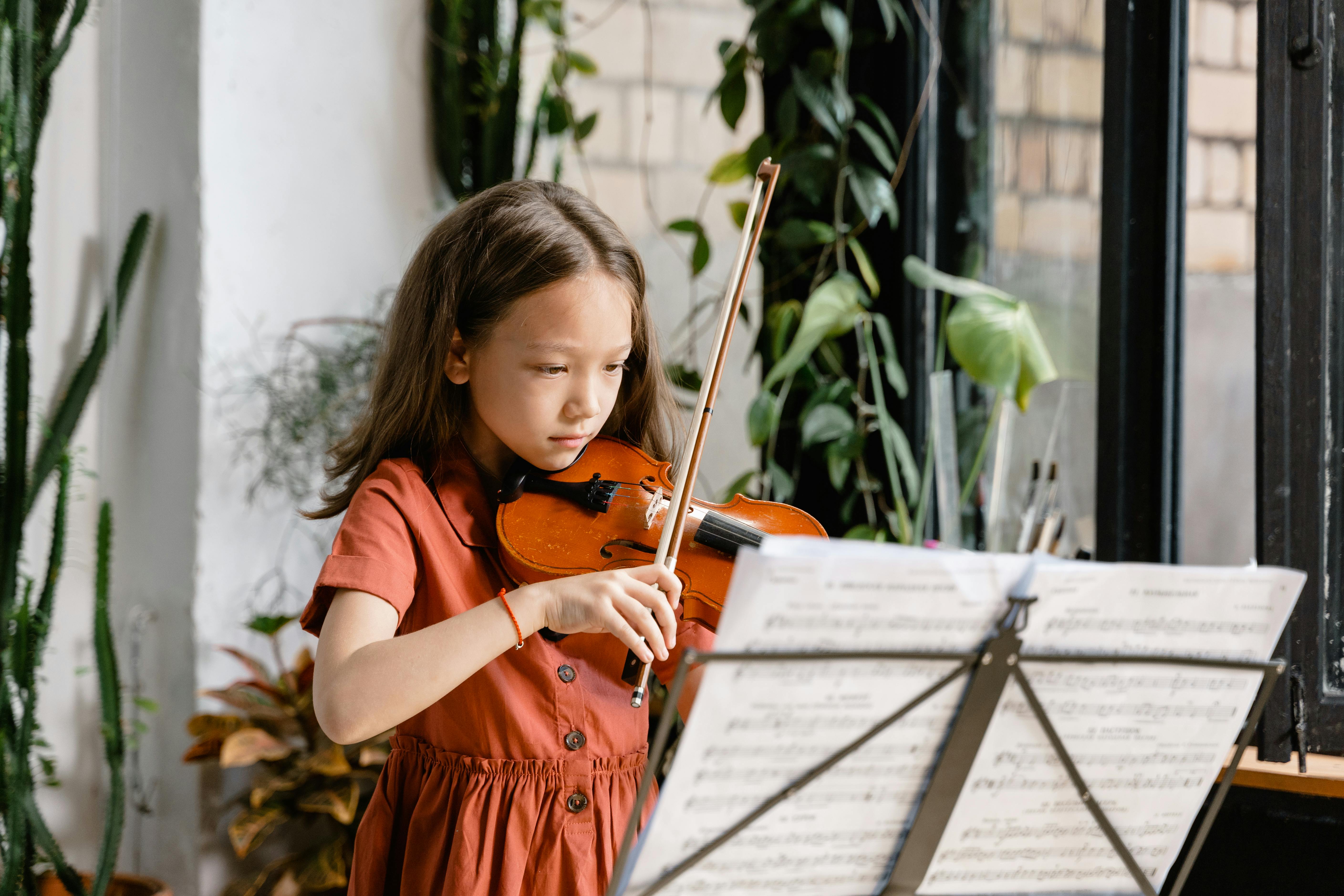 A young girl plays the violin with sheet music, focusing intently in an indoor setting.